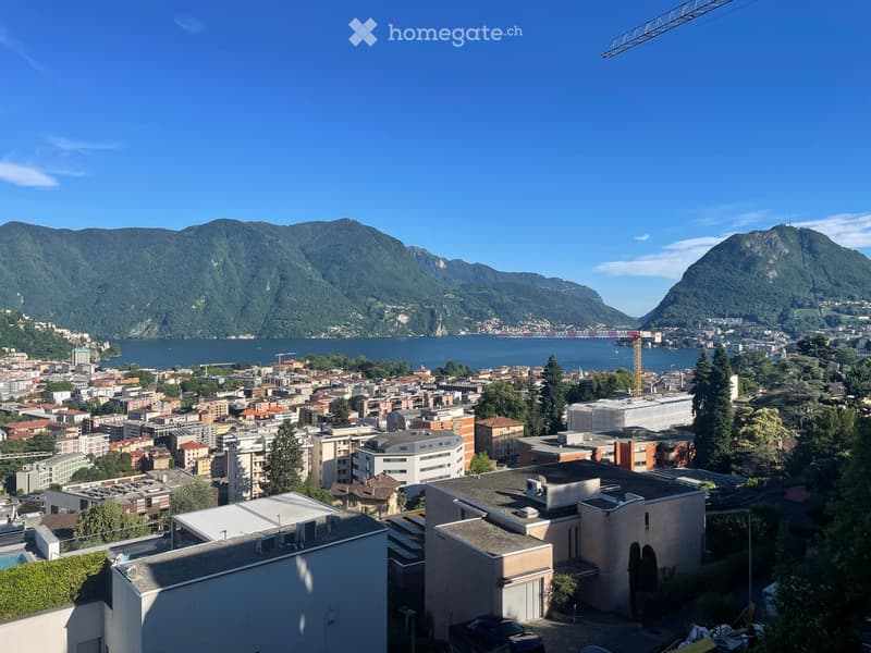 Aerial view of a cityscape with buildings, a lake, and surrounding mountains under a clear blue sky.