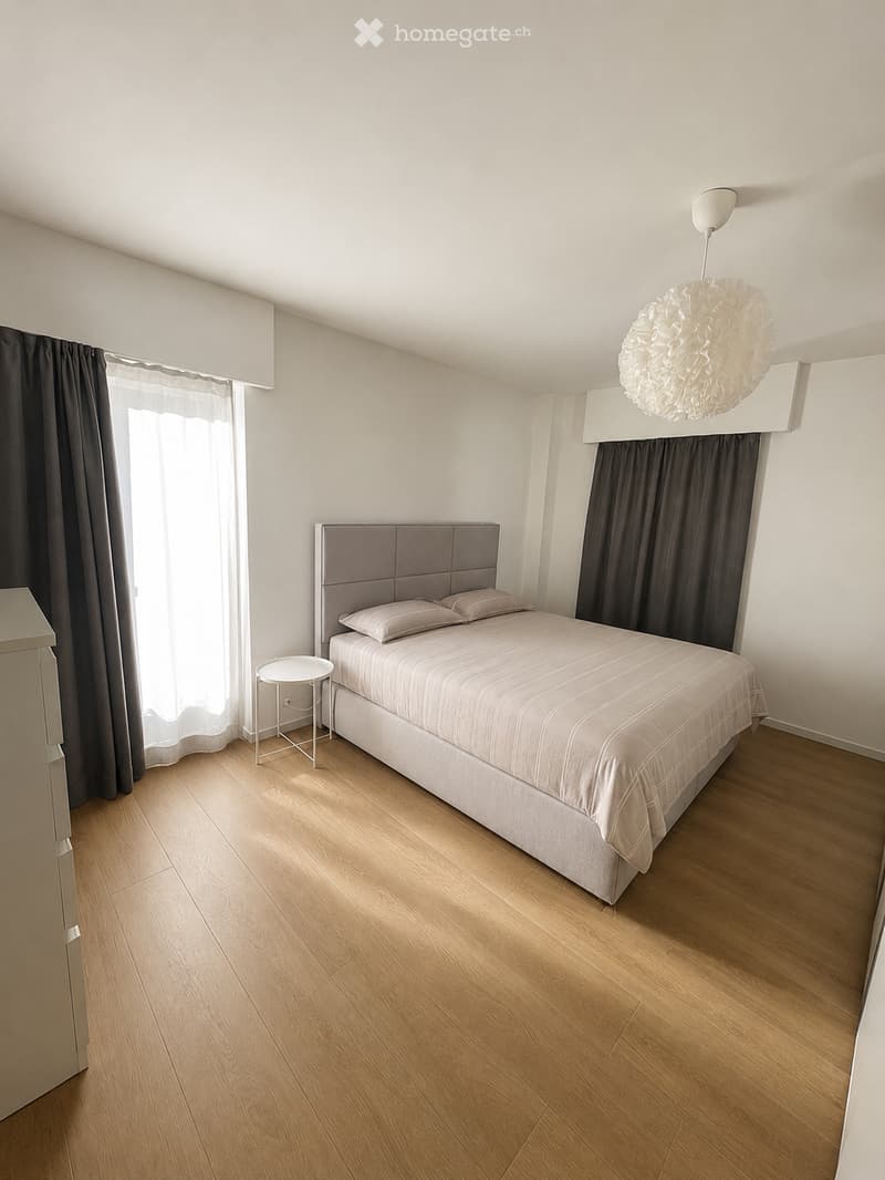 Modern bedroom with a double bed, nightstand, small round table, and large windows with dark curtains. Light wooden flooring and a decorative pendant light.