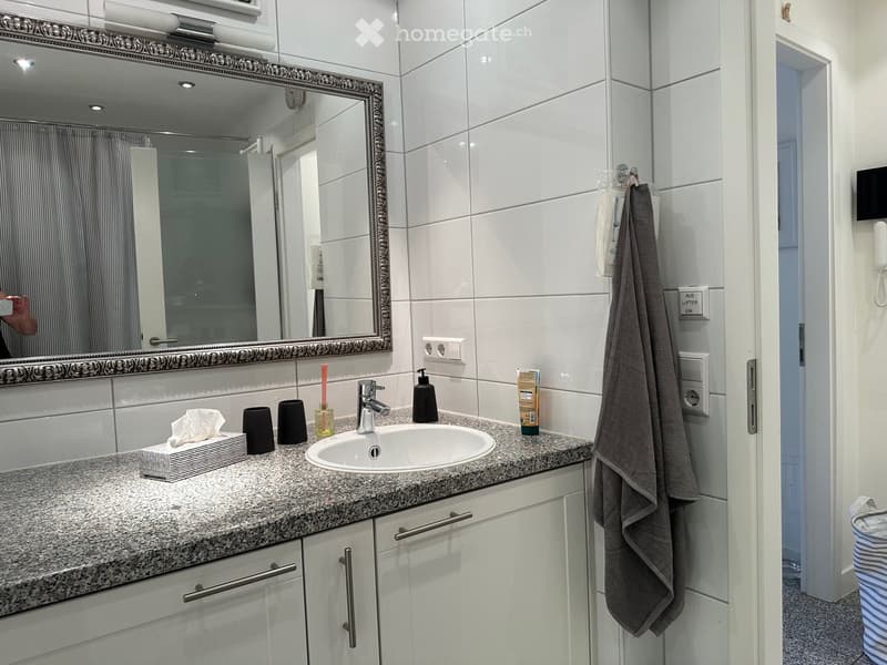 Modern bathroom with white tiled walls, granite countertop, single sink, mirror with ornate frame, towel, and small decorative items.