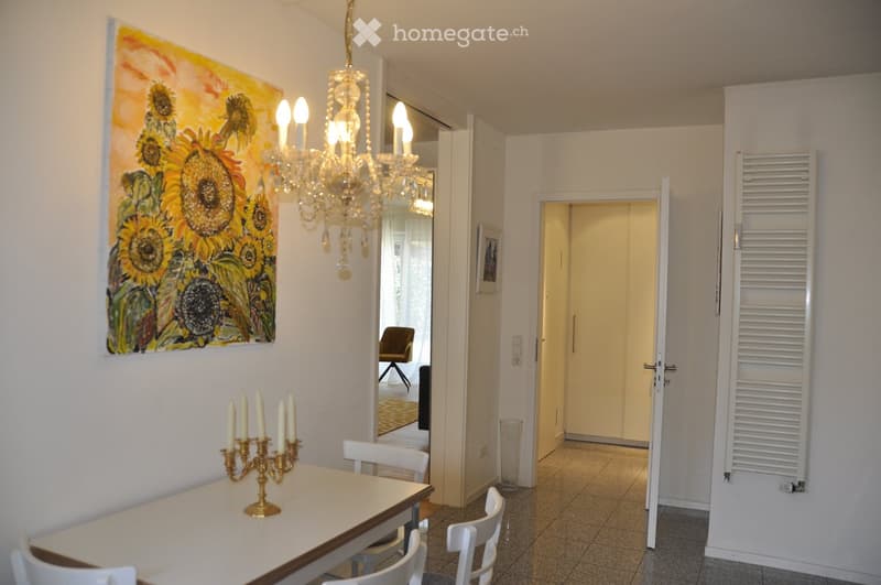 Dining area with a white table, four white chairs, a golden candelabra, a large mirror, and a radiator. A hallway leads to another room with a chair and a trash bin.