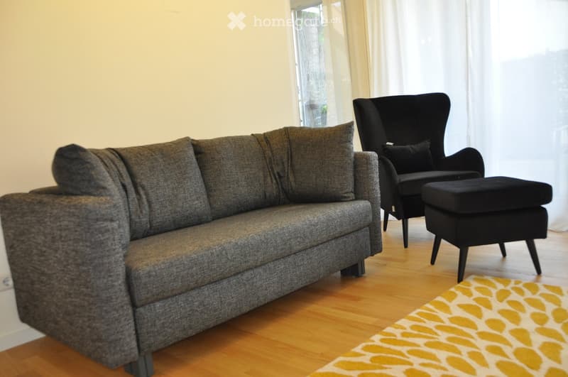 Living room with a grey sectional sofa, black armchair, ottoman, and a patterned rug on a wooden floor.
