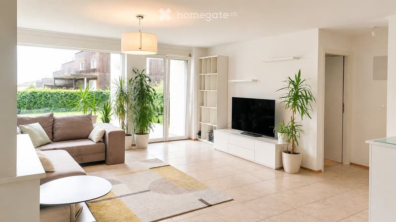 Bright living room with large sliding glass doors leading to a patio, a beige sectional sofa, a white coffee table, a flat-screen TV on a white entertainment unit, and several potted plants.