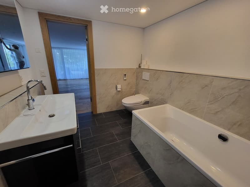 Modern bathroom with a white sink, chrome faucet, toilet, and bathtub, featuring beige tiled walls and dark tiled flooring.