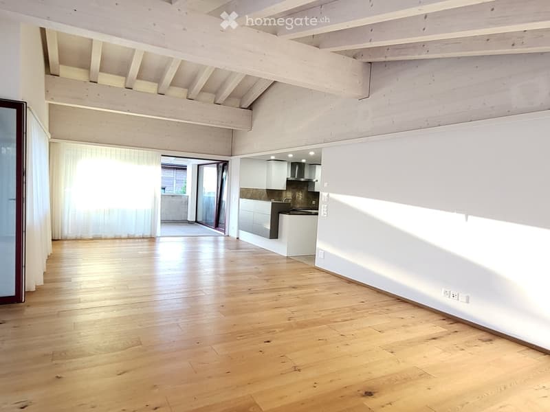 Open, unfurnished room with wooden flooring, white walls, and exposed wooden beams on the ceiling. Features a modern kitchen area with white cabinets and stainless steel appliances, and a large window with white curtains leading to a balcony.