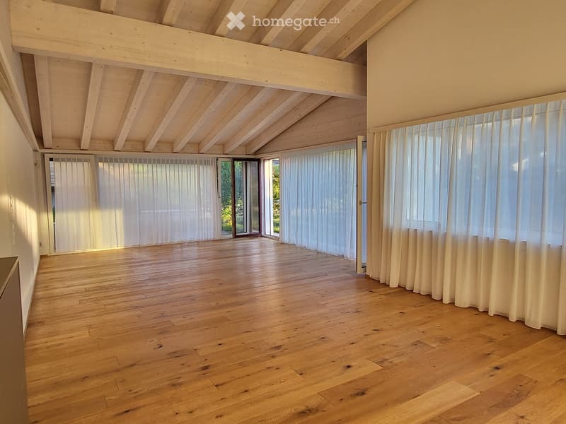 Bright, empty room with wooden flooring and large windows covered by white curtains, featuring exposed wooden beams on the ceiling.