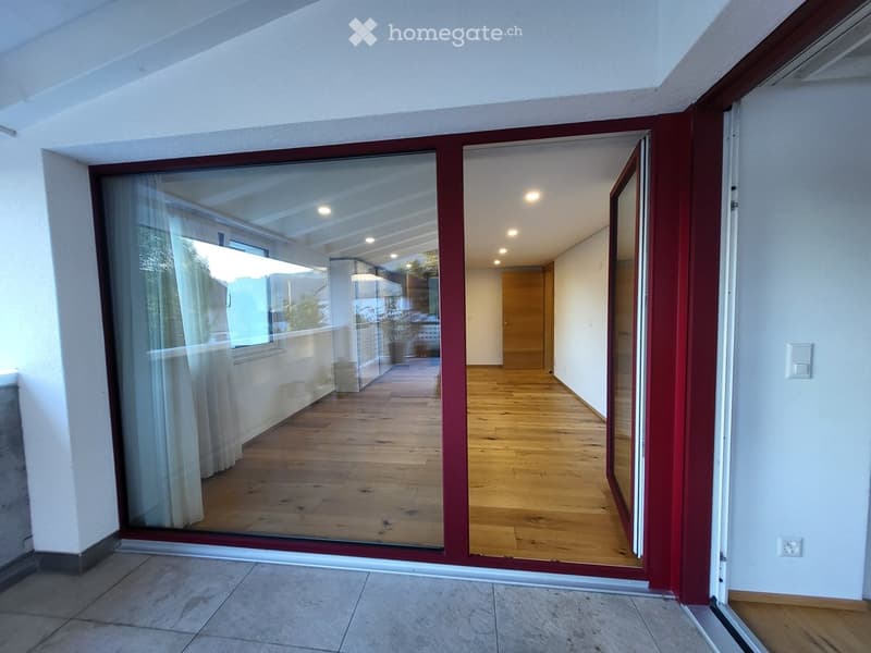 Open sliding glass doors lead from a tiled hallway into a wooden-floored room with white walls and recessed lighting.