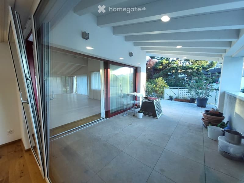 Spacious balcony with tiled floor, white walls, and a covered area. Visible are potted plants, a small table, and a pet carrier.
