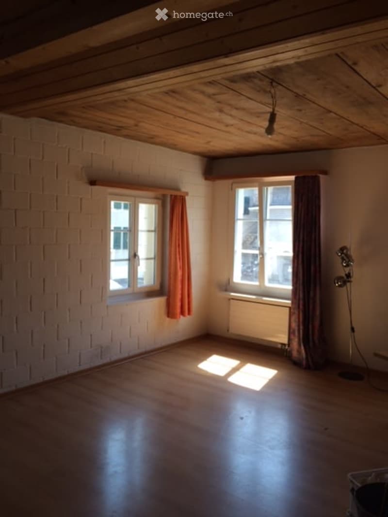 Empty room with wooden flooring, white brick walls, two windows with curtains, and a radiator. No furniture or equipment present.