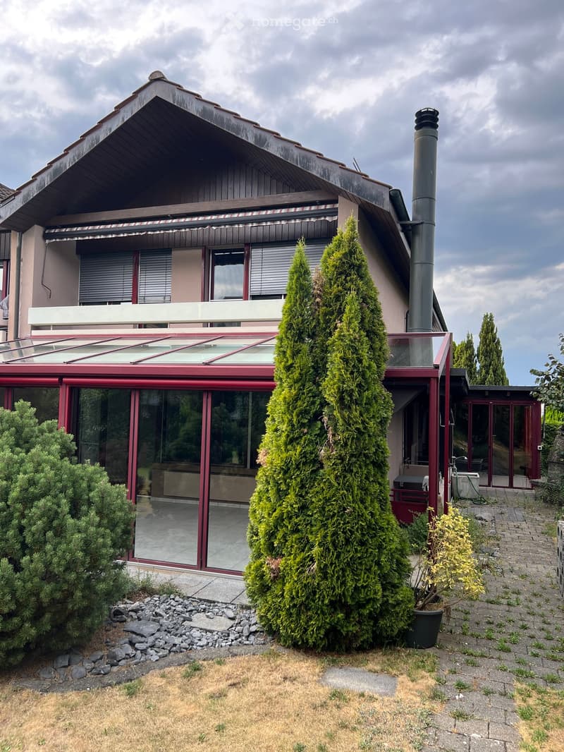 Two-story house with a modern design, featuring a large glass-enclosed patio area, a chimney, and surrounding greenery including tall coniferous trees and shrubs.