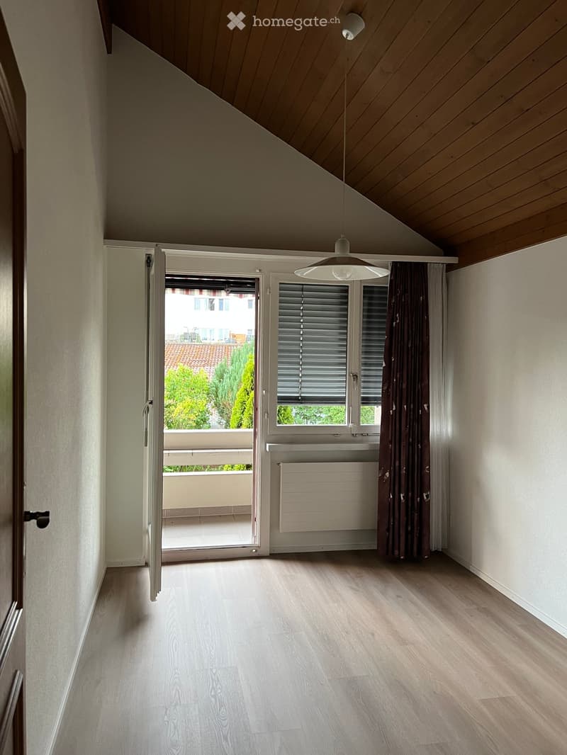 Empty room with wooden flooring, white walls, and a large window with blinds. An open sliding door leads to a balcony with outdoor view.