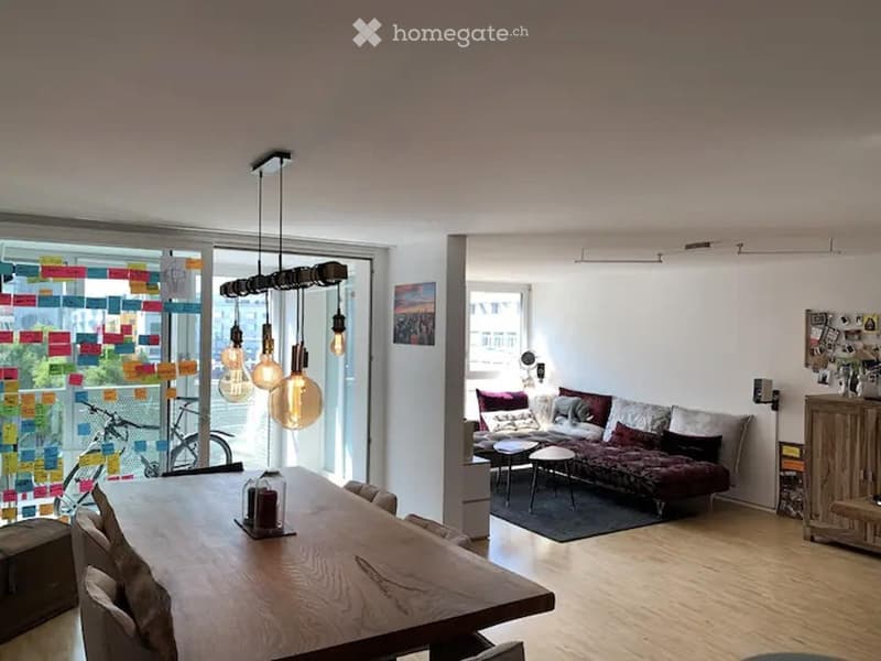 Open-plan living and dining area with a wooden dining table, chairs, a sofa with pillows, a small stool, and a wooden cabinet. Large windows and pendant lights are visible.