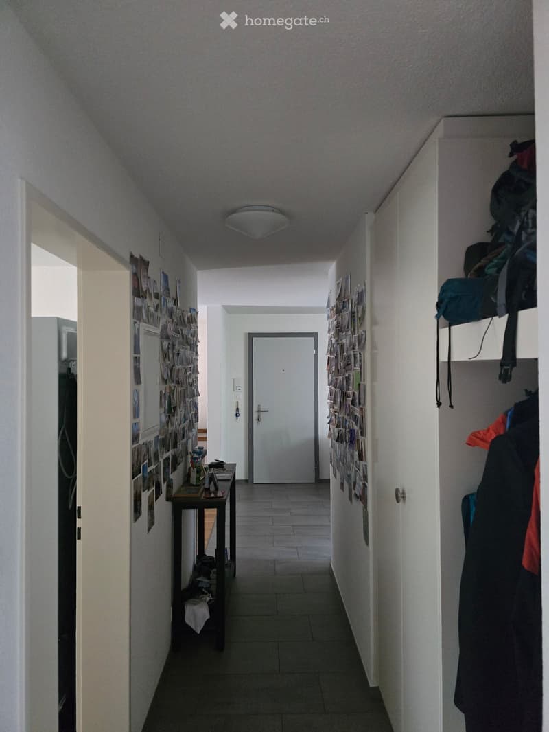A narrow hallway with white walls, tiled floor, and a ceiling light. Photos are pinned to the walls, and there is a small table with items on it to the left. A closet with clothes and bags is visible on the right, and a closed door is at the end of the ha