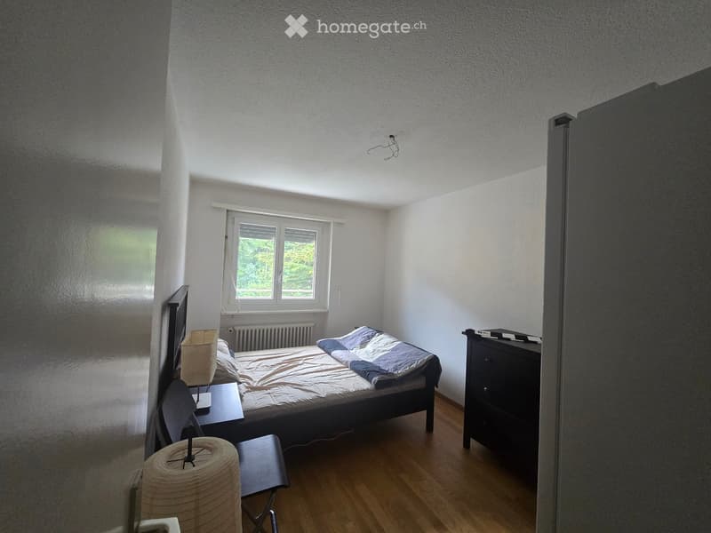 A small, well-lit bedroom with a bed, nightstand, lamp, chair, and a drawer. The room features wooden flooring and large windows with blinds.