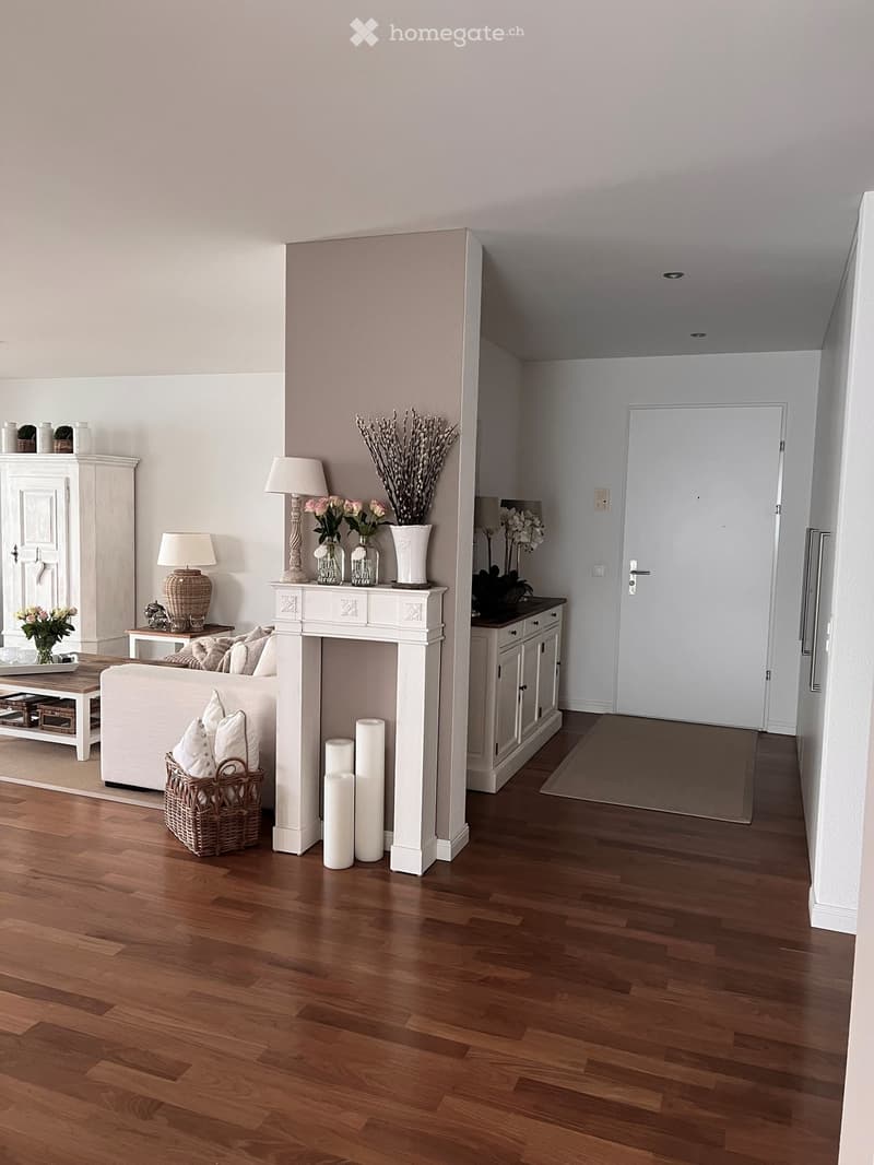A well-lit living room with a white fireplace, a sofa, coffee table, table lamps, and decorative vases. The room features wooden flooring and a hallway leading to another area.
