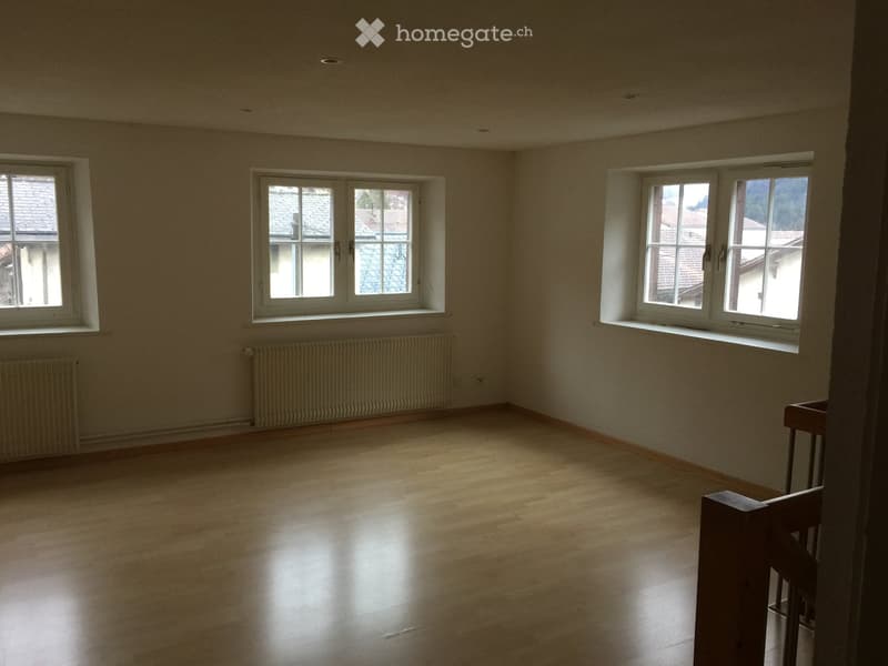 Empty room with wooden flooring, white walls, and multiple windows offering views of rooftops. A radiator is visible along one wall, and part of a wooden staircase railing is seen in the foreground.