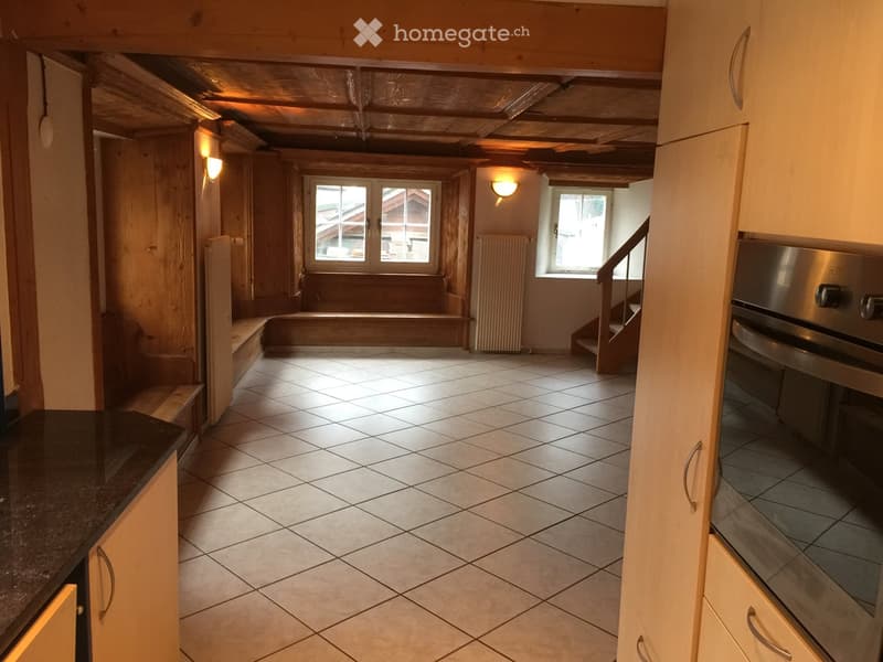 A cozy, rustic kitchen area with wooden beams, tiled floor, and natural light from two windows. Features a countertop, built-in oven, radiator, and a staircase leading upwards.