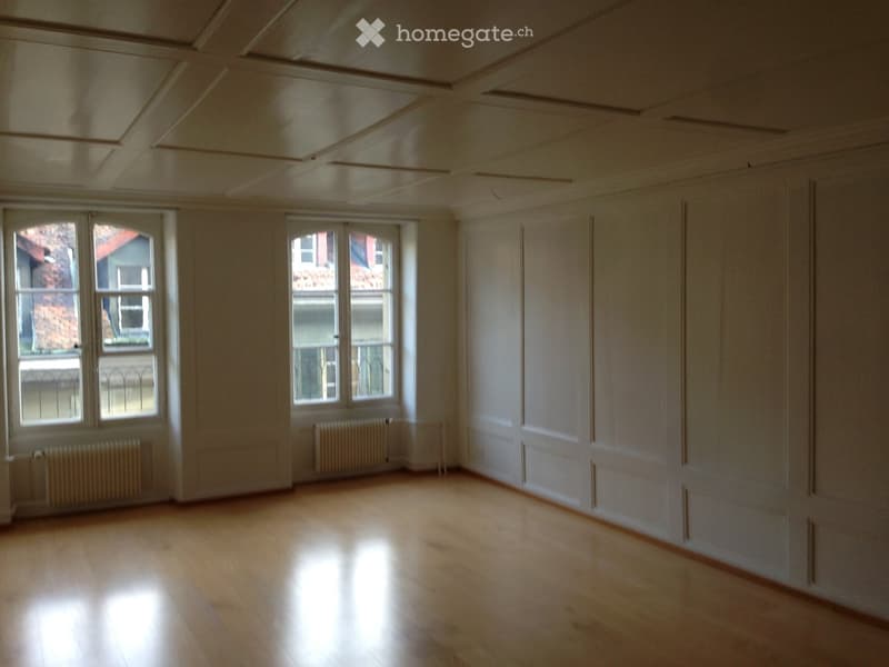 Empty room with wooden flooring, white walls, and large windows with radiators beneath them.