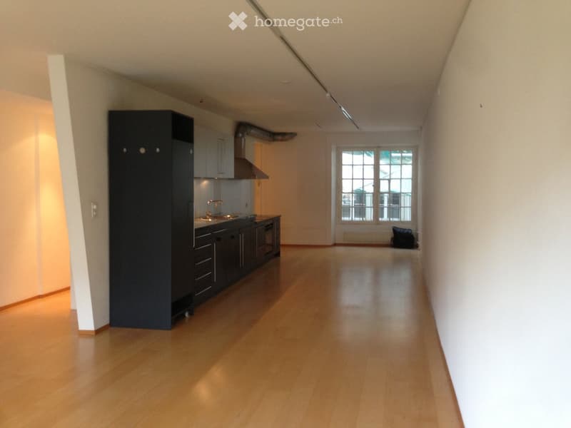 Empty room with wooden flooring, white walls, and a small kitchen area with cabinets and a sink visible in the background.