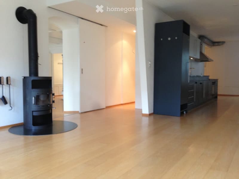 Open-plan space with a modern fireplace and fitted kitchen cabinets, featuring light wooden flooring and white walls.