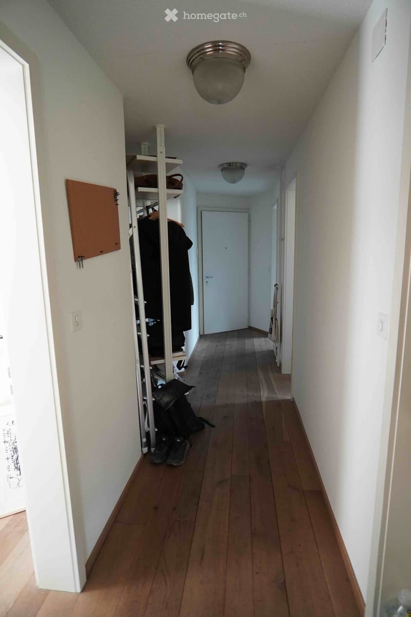 A narrow hallway with wooden flooring, white walls, and a ceiling light fixture. A coat rack with jackets and shoes is visible on the left side, and a closed door is at the end of the hallway.