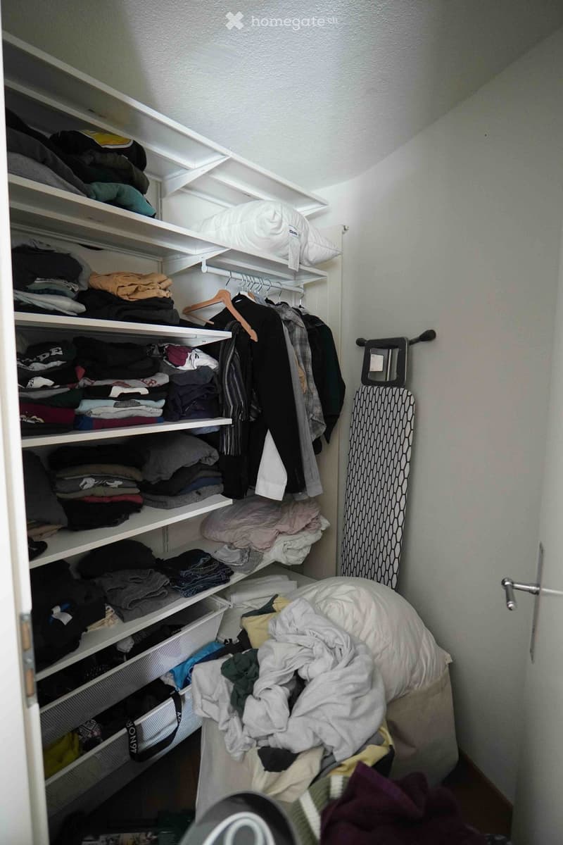 A well-organized closet with multiple shelves filled with folded clothes, a hanging rod with garments, a pillow on top, and an ironing board leaning against the wall.