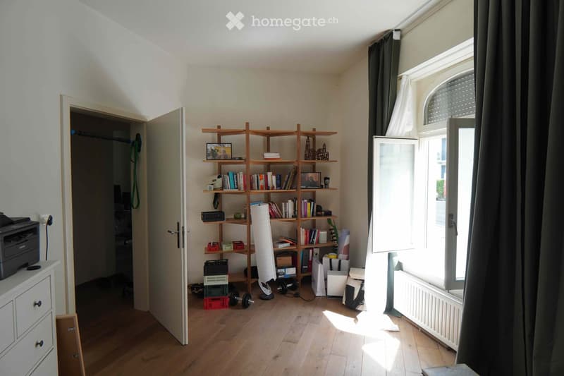 A well-lit room with wooden flooring, a large open shelving unit filled with books and decorative items, a printer on a small cabinet, and gym equipment near the window.