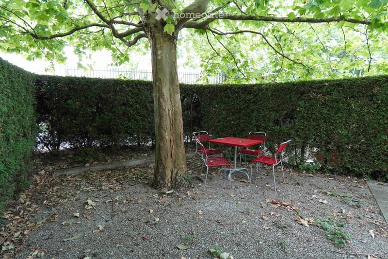 A small outdoor seating area with a red metal table and four matching chairs, surrounded by a gravel ground, a large tree, and a neatly trimmed hedge wall.