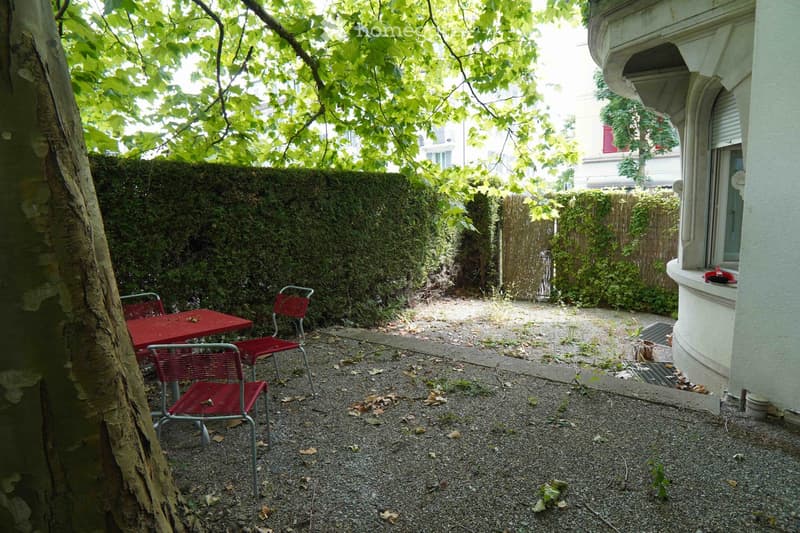 Small outdoor garden area with a red table and chairs, surrounded by greenery and a tree, with a building in the background.