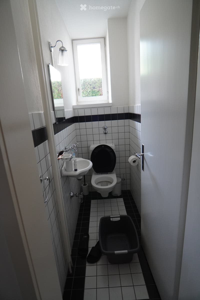 Small bathroom with white tiled walls, black accents, toilet, sink, mirror, and a window letting in natural light.