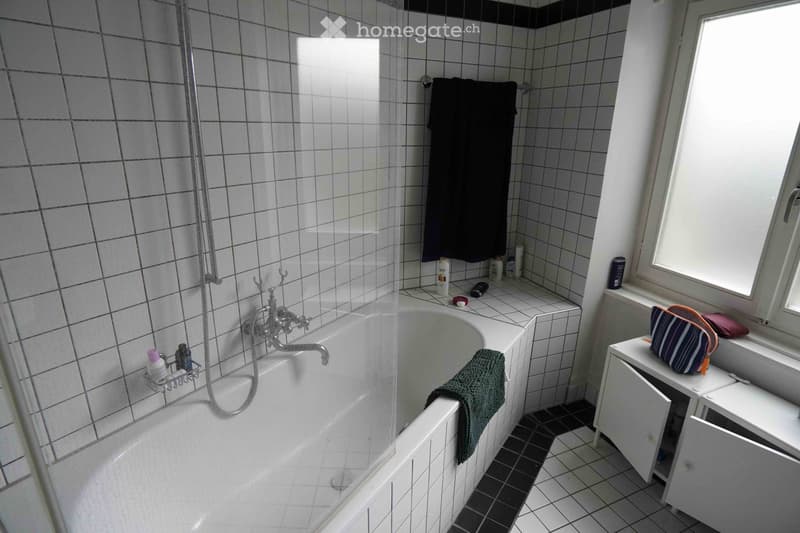 Bathroom with a bathtub, tiled walls, a towel hanging on the side, a small cabinet with a handbag on top, and various toiletries on the windowsill.