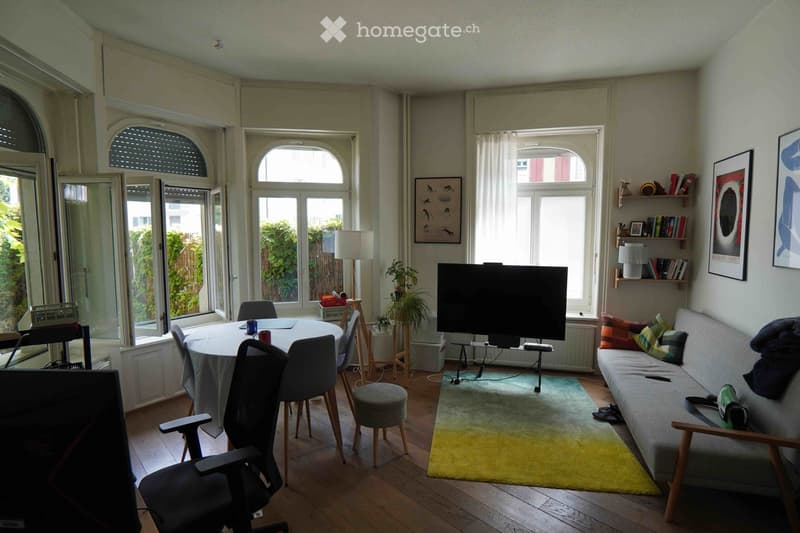 Bright living room with large windows, wooden flooring, a sofa with colorful cushions, a coffee table, a dining table with chairs, a TV, a floor lamp, and a small plant on a side table.