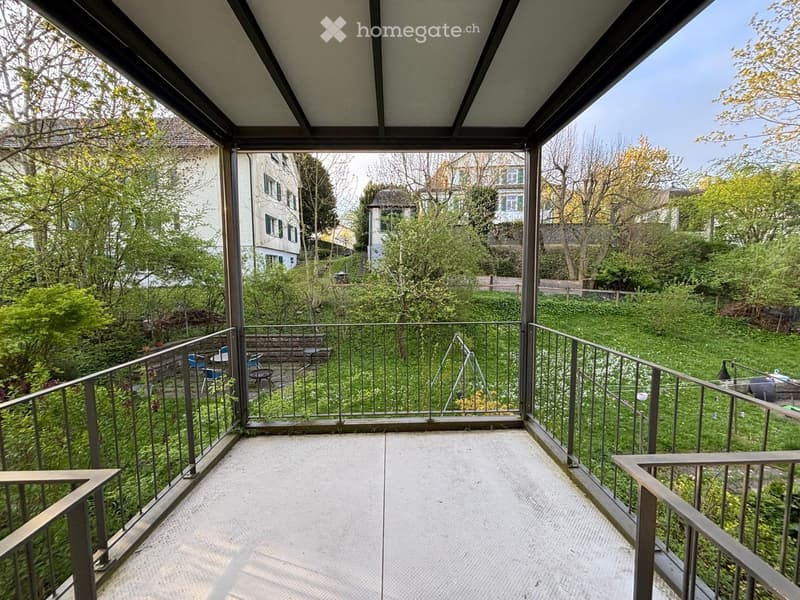Balcony / Terrace. Balcony with metal railings, concrete floor, overlooking a green garden area with trees, chairs, and a small table.
