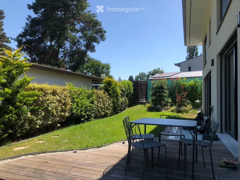 Wooden deck with a table and chairs, surrounded by a well-maintained lawn and various trees and shrubs.