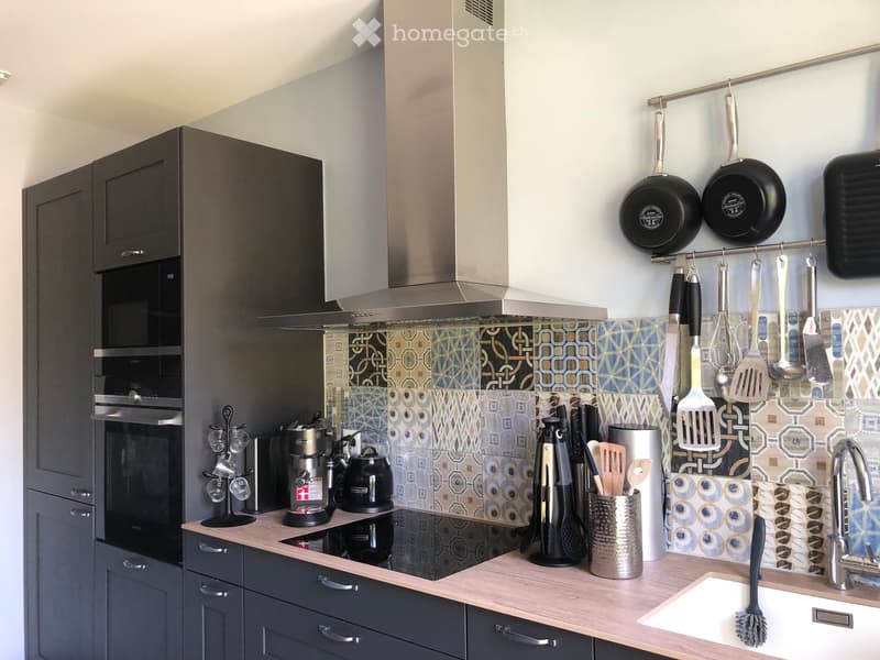 Modern kitchen with grey cabinets, stainless steel appliances, and a decorative backsplash. Features include a coffee maker, teapot, hanging pans, and a sink with a modern faucet.