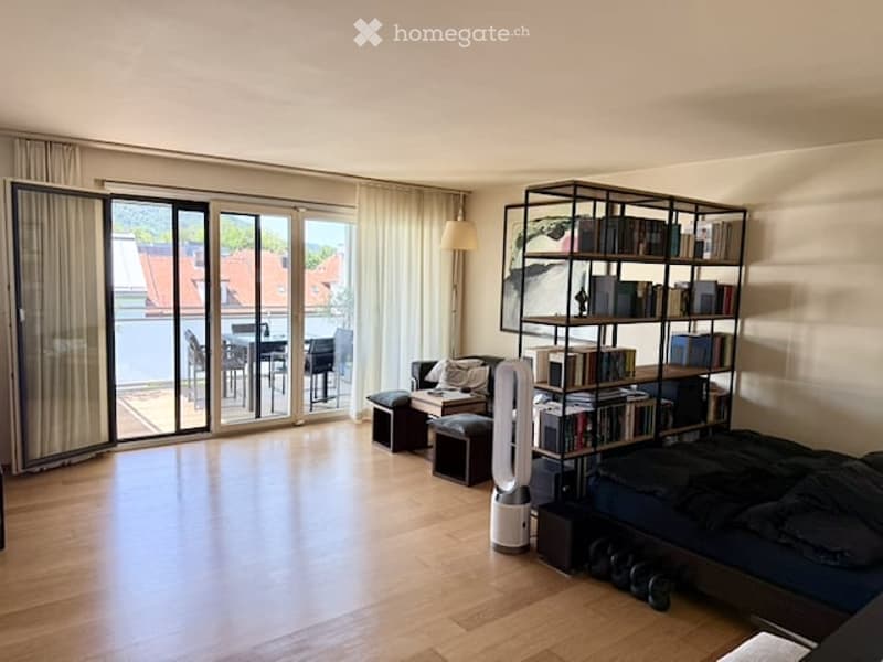 Bright, open living space with wooden flooring, large glass doors leading to a balcony, a sofa, a bookshelf, a fan, and a bed.