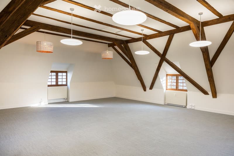Empty room with wooden beams ceiling, two windows, white walls, and white carpet.