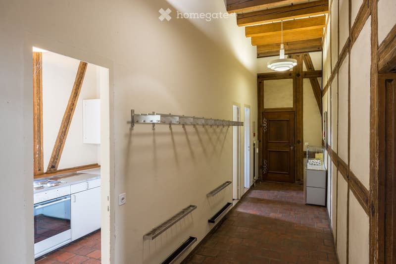 A hallway, white walls, wooden ceiling, kitchen on left, hallway lamp, closed wooden door