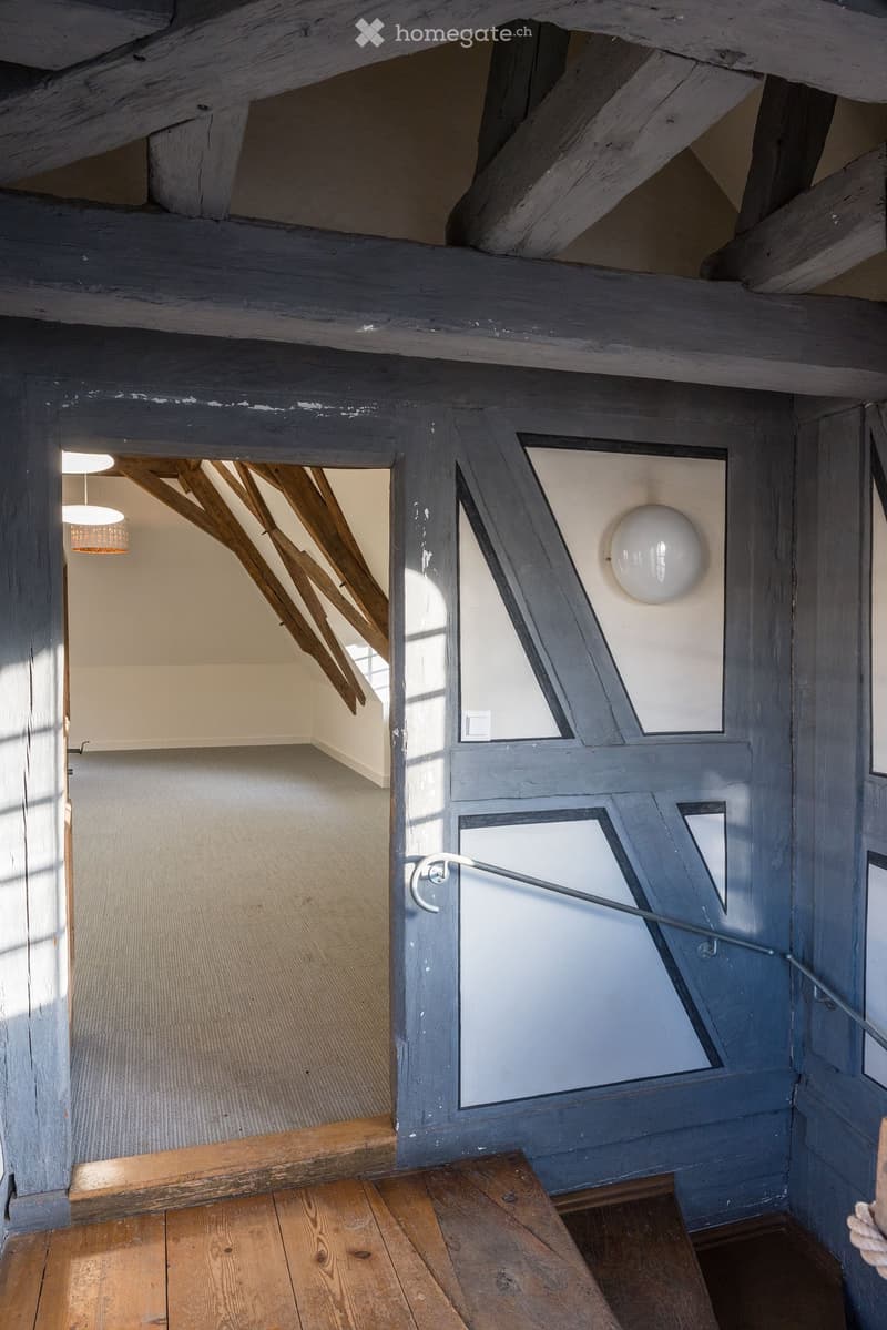 Room with wooden floor, wooden beams, white walls, open door, small window, carpet, chandelier, wall lamp