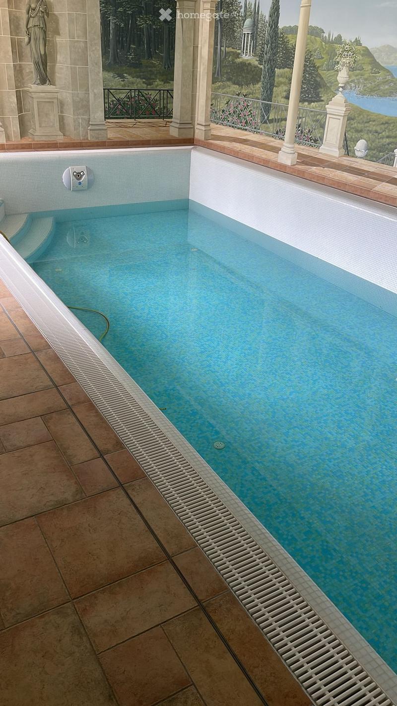 A rectangular swimming pool with clear blue water, surrounded by a tiled deck and a white drainage grate along one edge. The pool is situated in an outdoor area with a scenic view of a landscape and sea beyond.