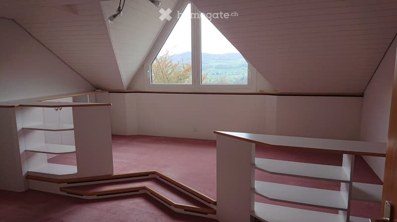 An empty attic room with wooden stairs, built-in shelving, and skylight windows offering a scenic view.