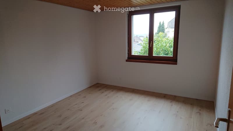 Empty room with light wooden flooring and white walls, featuring a large window with wooden frame and view of greenery outside.