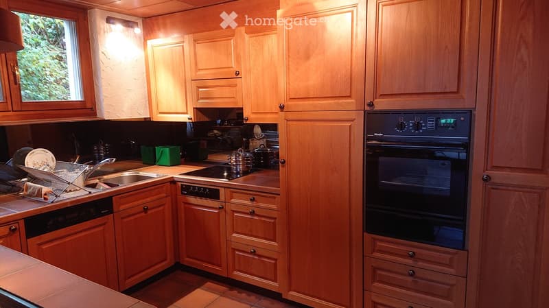 A well-lit kitchen with light wooden cabinets, stainless steel appliances, and a window overlooking greenery. The kitchen features a sink, stove, oven, and dishwasher, with various kitchen items on the counter.