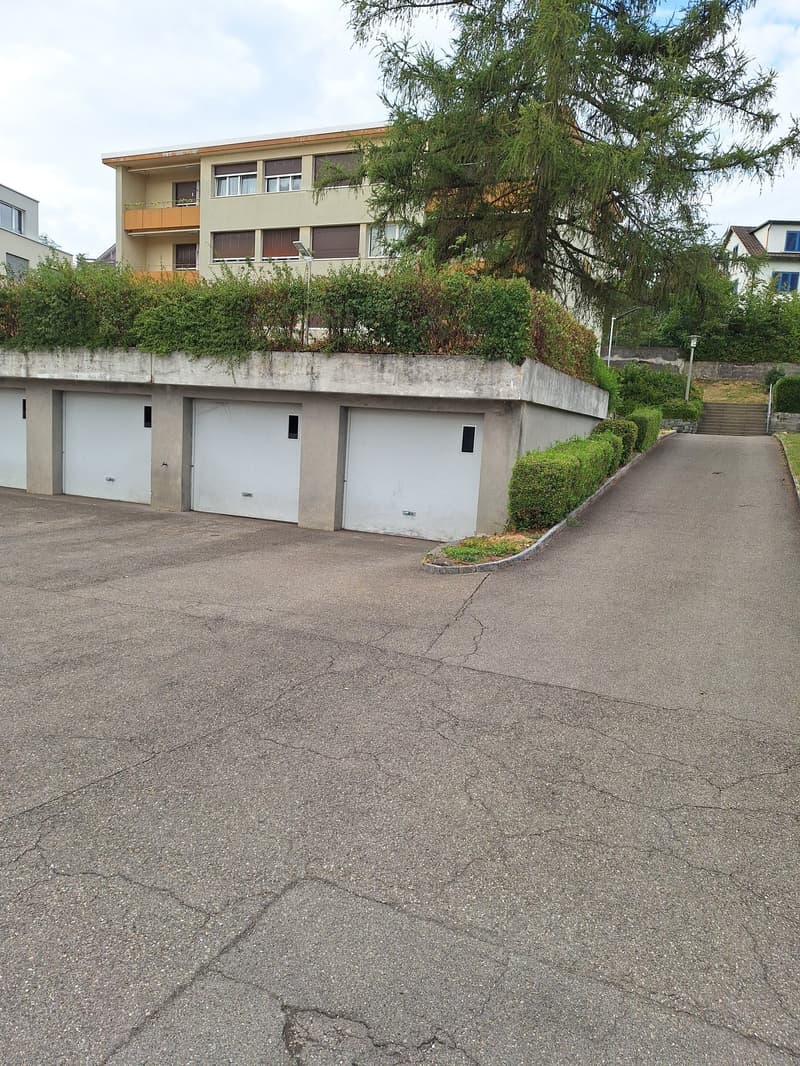 A modern two-story building with a flat roof, multiple windows, and a large garage area. The building is surrounded by greenery and has a concrete driveway leading to the entrance.
