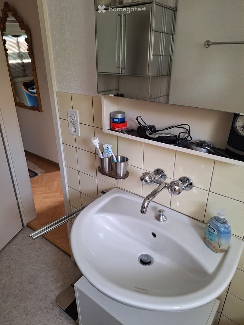 Modern bathroom with a white sink, chrome faucet, toothbrush holder, soap dispenser, and a mirrored cabinet above. The sink has a sleek, curved design with a built-in drain and overflow hole.
