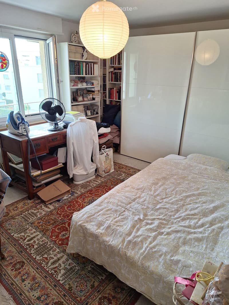 A well-lit bedroom with a large bed, patterned bedspread, bookshelf, fan, and a wooden table with various items on it.