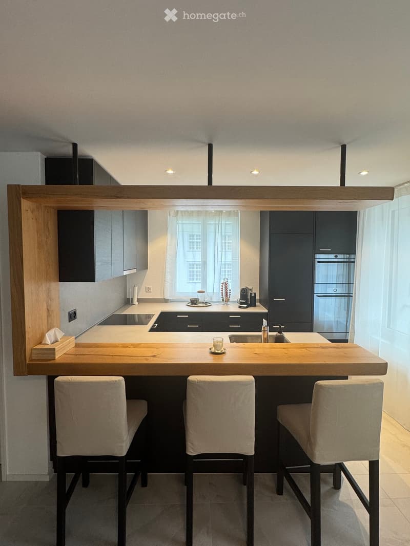 Modern kitchen with wooden counter, black cabinets, white tiled floor, island with bar stools, sink, and faucet, built-in oven, and microwave