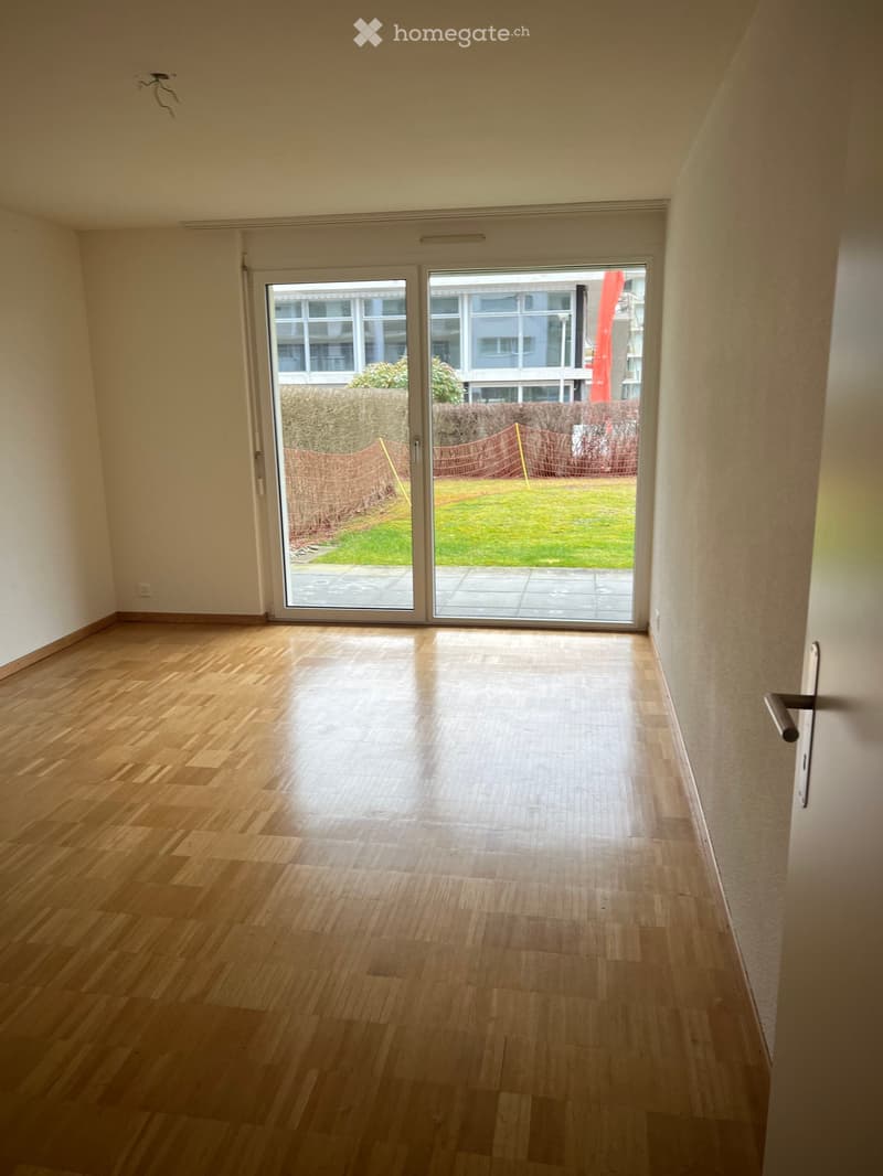 Empty room with wooden flooring, large sliding glass doors leading to a patio and garden area, white walls, and a visible electrical outlet.