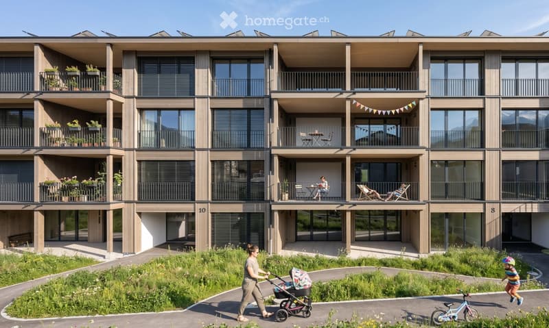 Modern multi-story residential building with balconies, large windows, and a landscaped ground area with greenery and pathways.