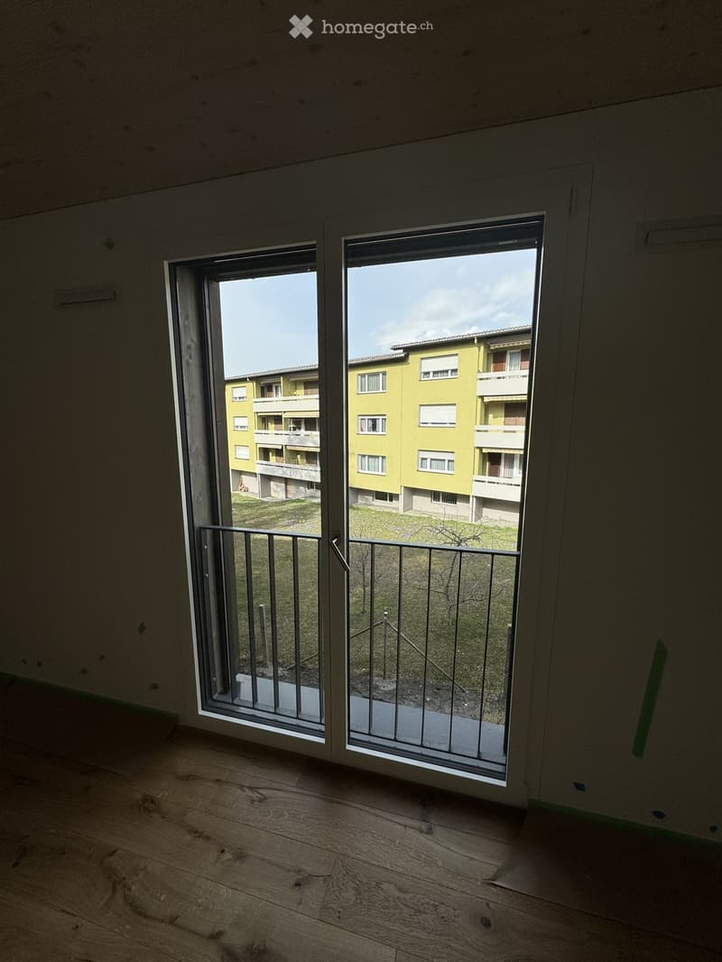 Empty room with wooden flooring and large sliding glass doors leading to a balcony overlooking a residential area.