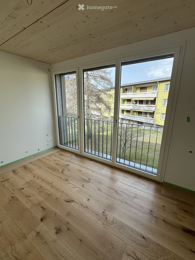 Empty room with wooden flooring and large sliding glass doors leading to a balcony with metal railings. The walls are painted white, and the ceiling has exposed wooden beams.